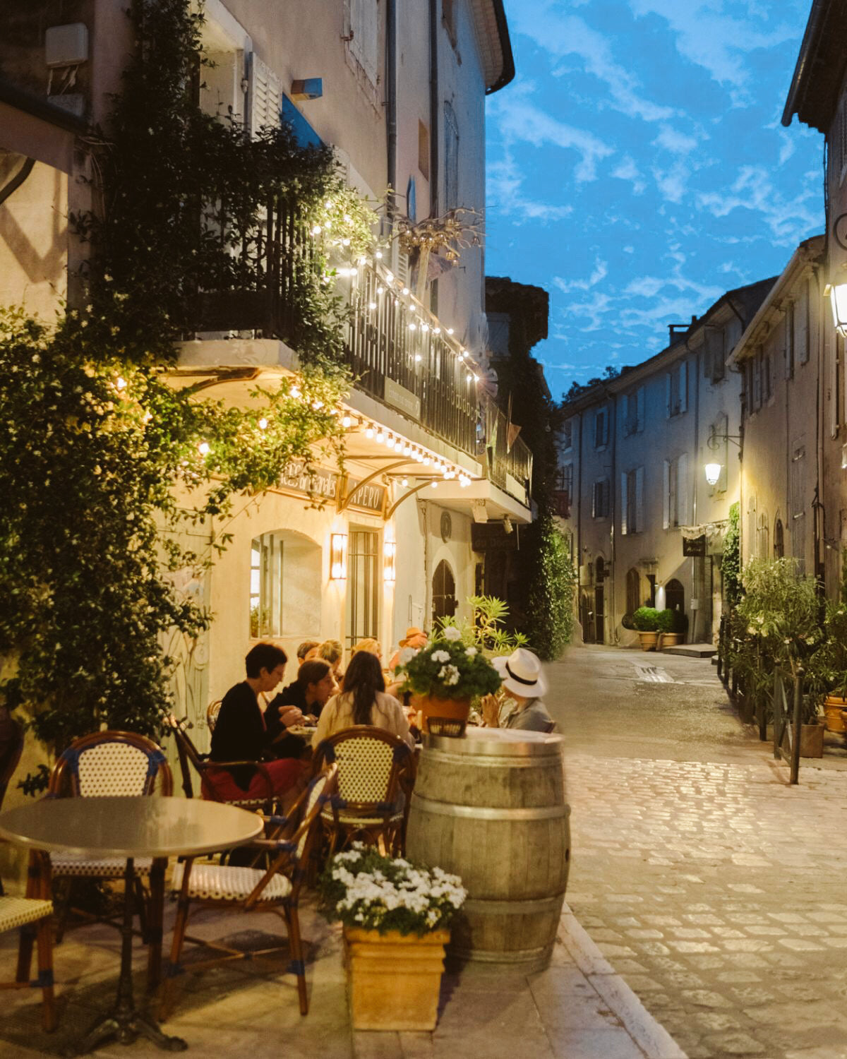 Cobblestone street in Lourmarin at night, with climbing plants, pastel toned shuttered windows and a lively restaurant with outdoor seating.