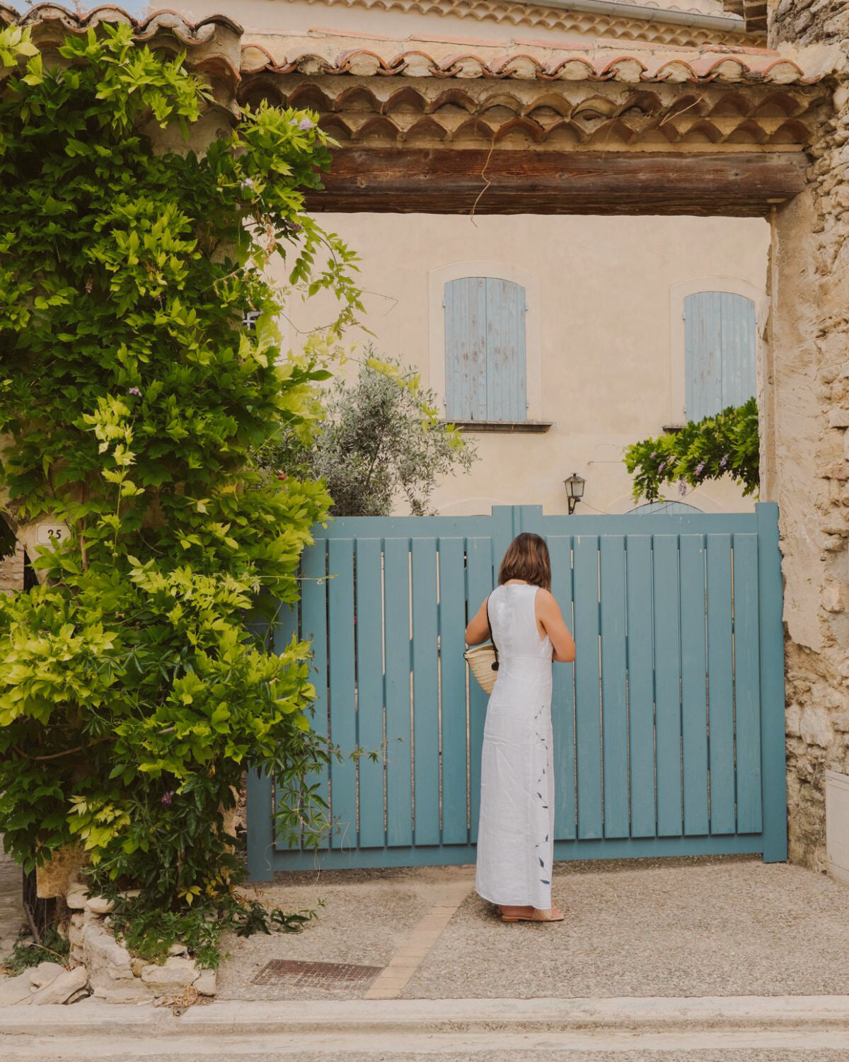 Woman wearing a white linen dress, in front of a charming blue gate in Venasque. Shuttered blue windows and climbing plants.