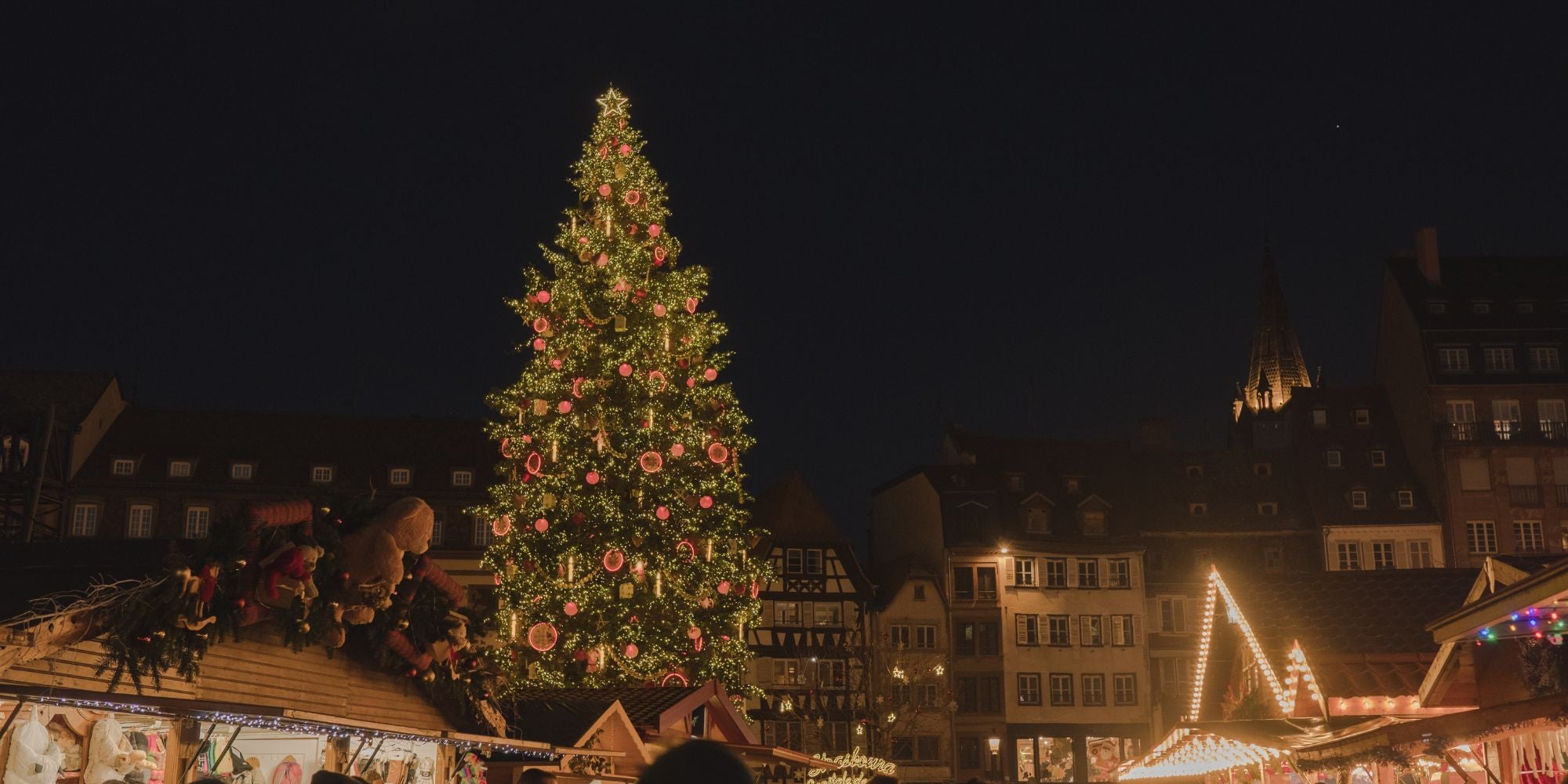 Christmas market setting at night, in the streets of Strasbourg. A high tree with ornaments and lights, a crowded market and buildings in the back.
