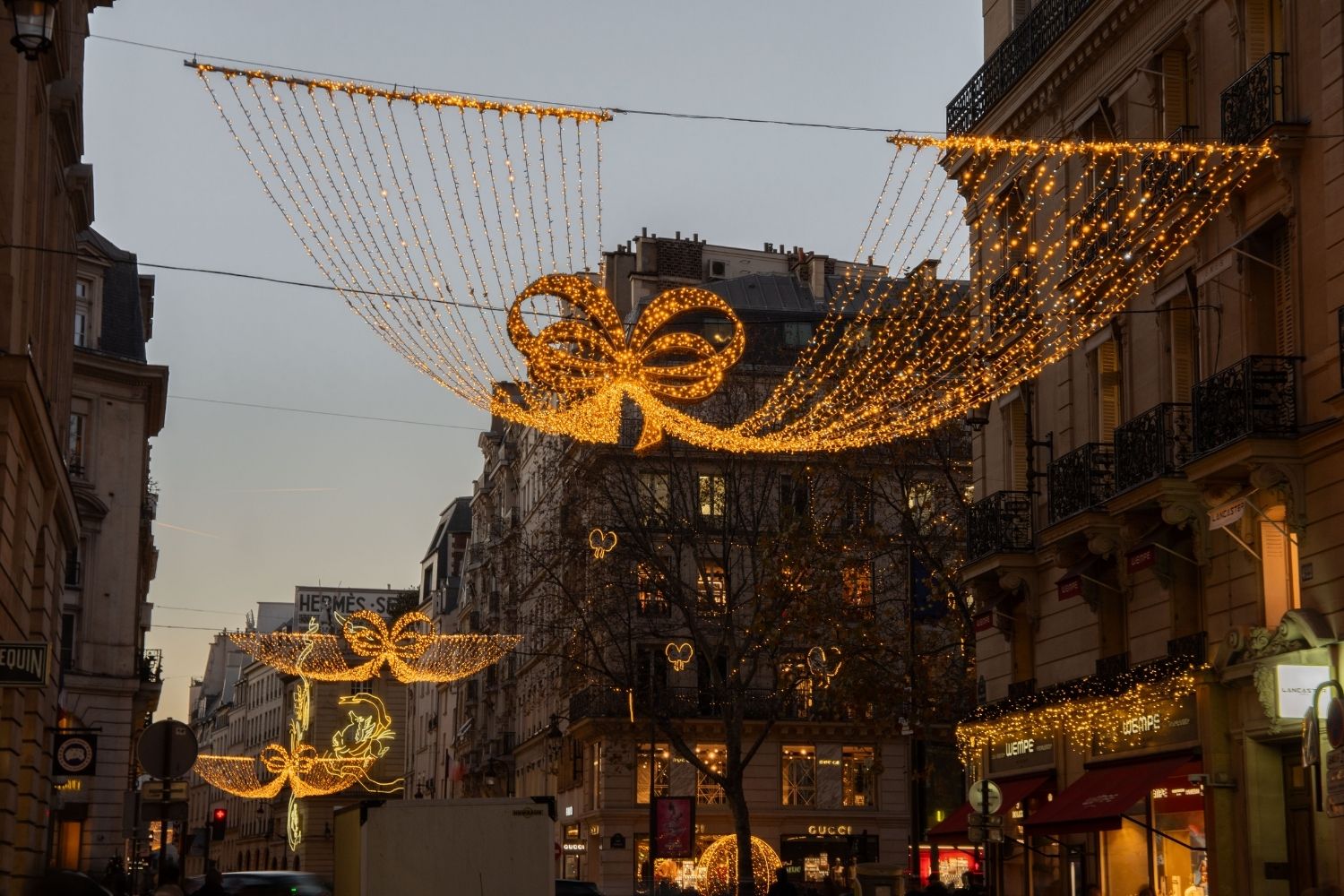 Paris street at sunset, with classic buildings where cars pass through. Christmas ornaments adorn the streets, and the doors and windows of the buildings.