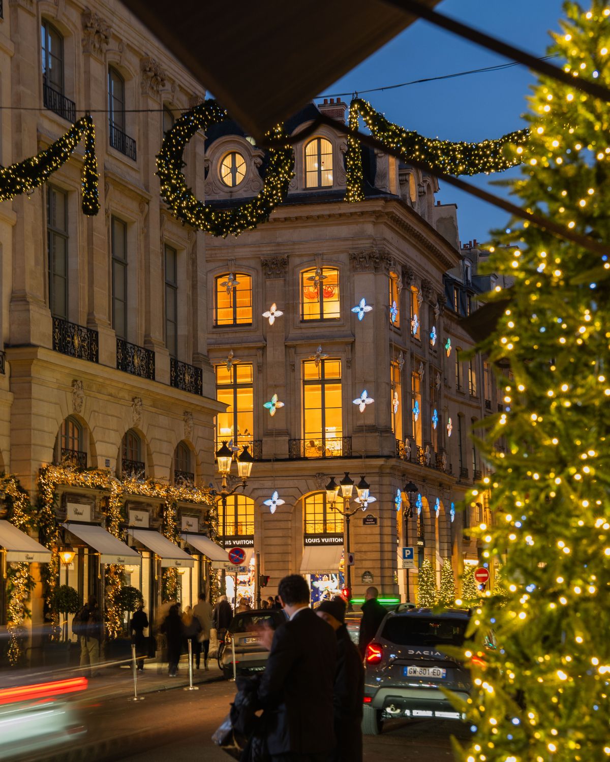 Paris street at sunset, with classic buildings where cars pass through. Christmas ornaments adorn the streets, and the doors and windows of the buildings.
