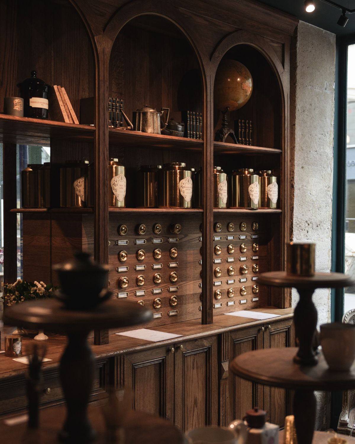 A wooden shelf, with golden brass pots, buttons on the back and decorative accessories of the top shelf, like candles, books, a black pot, a small lamp and world globe.