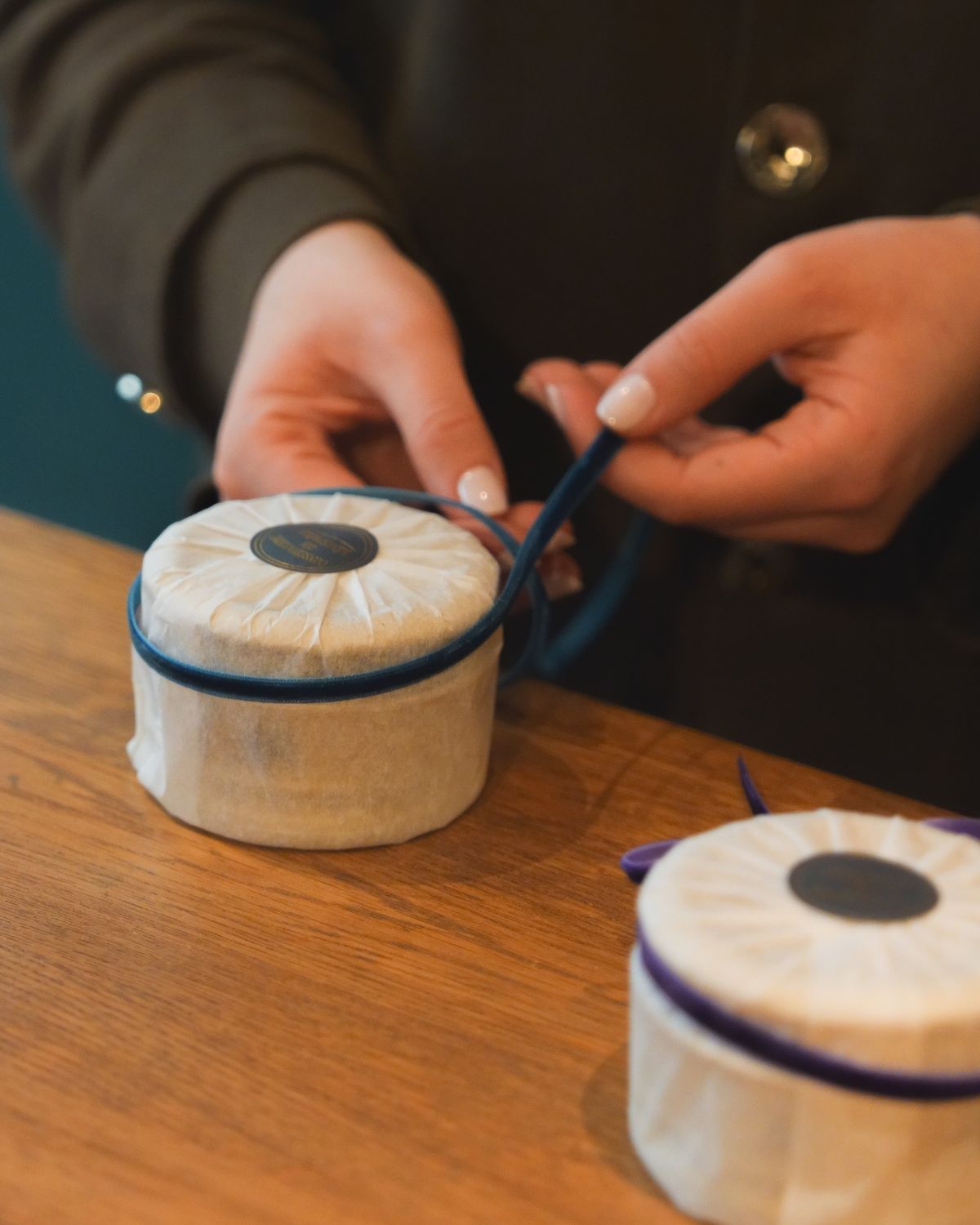 A lady's hands wrapping a circular product, with a blue velvet ribbon, on top of a cherry wooden counter.
