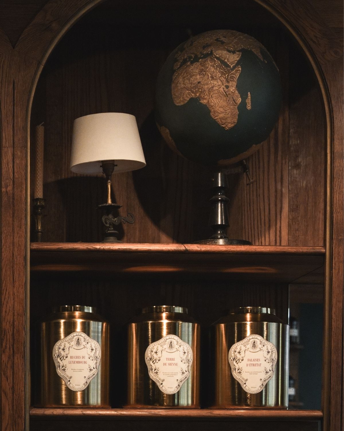 A wooden shelf, with golden brass pots, and decorative accessories of the top shelf, like a candle, a small lamp and world globe.