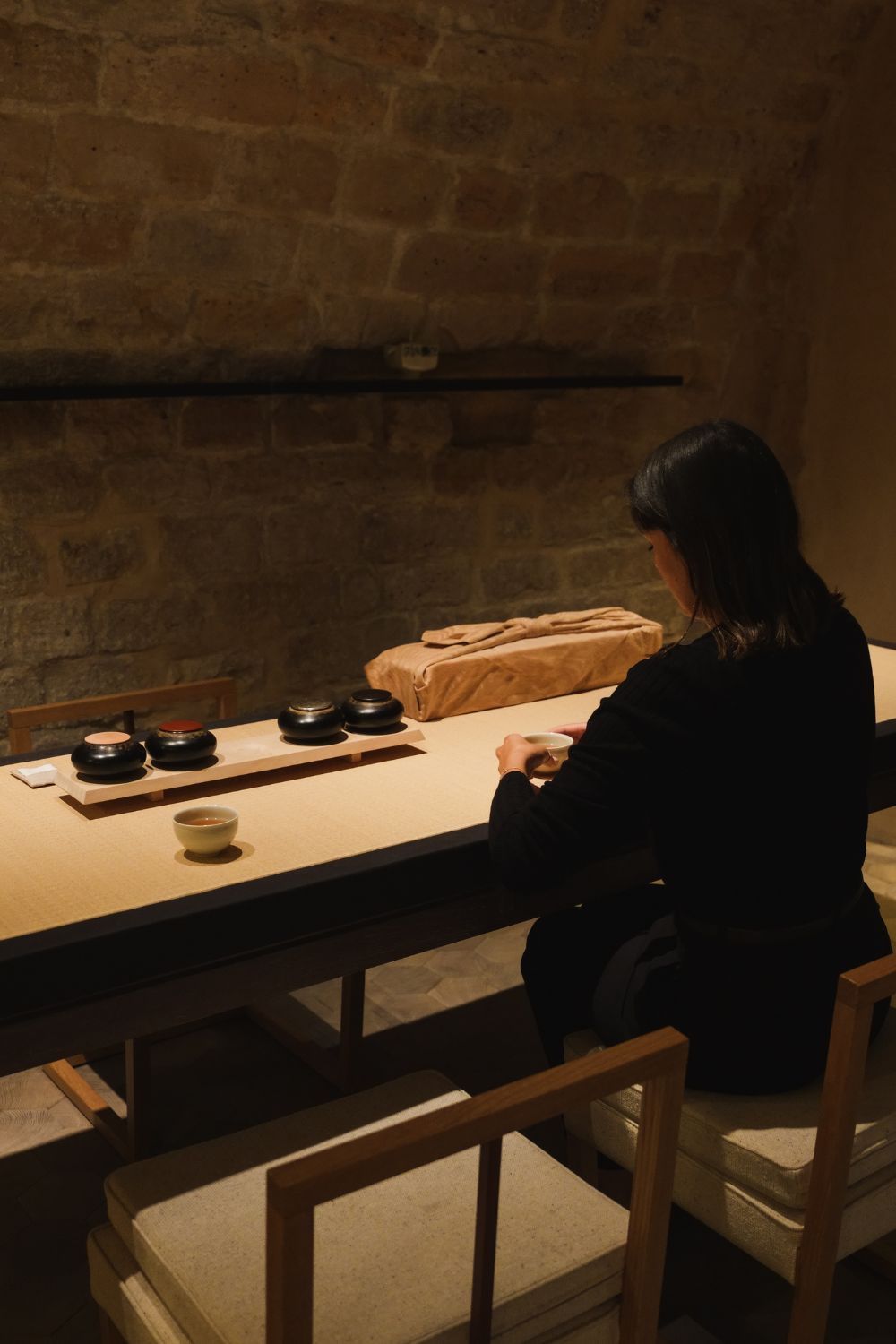 A lady in black, sited facing a table, while drinking tea, inside a tea room with light fabric and wooden furniture and stone walls.