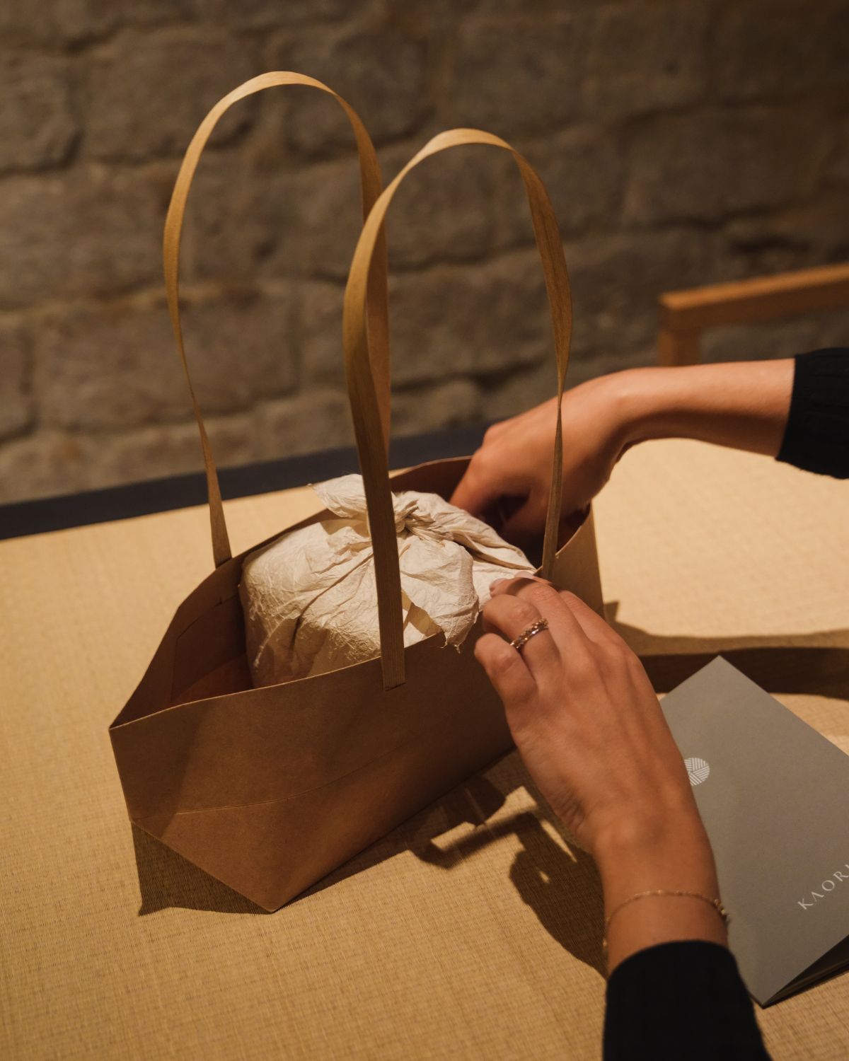 A paper bag with a wrapped product on the inside, and a ladies hand opening it on top of a light table, next to a stone wall.