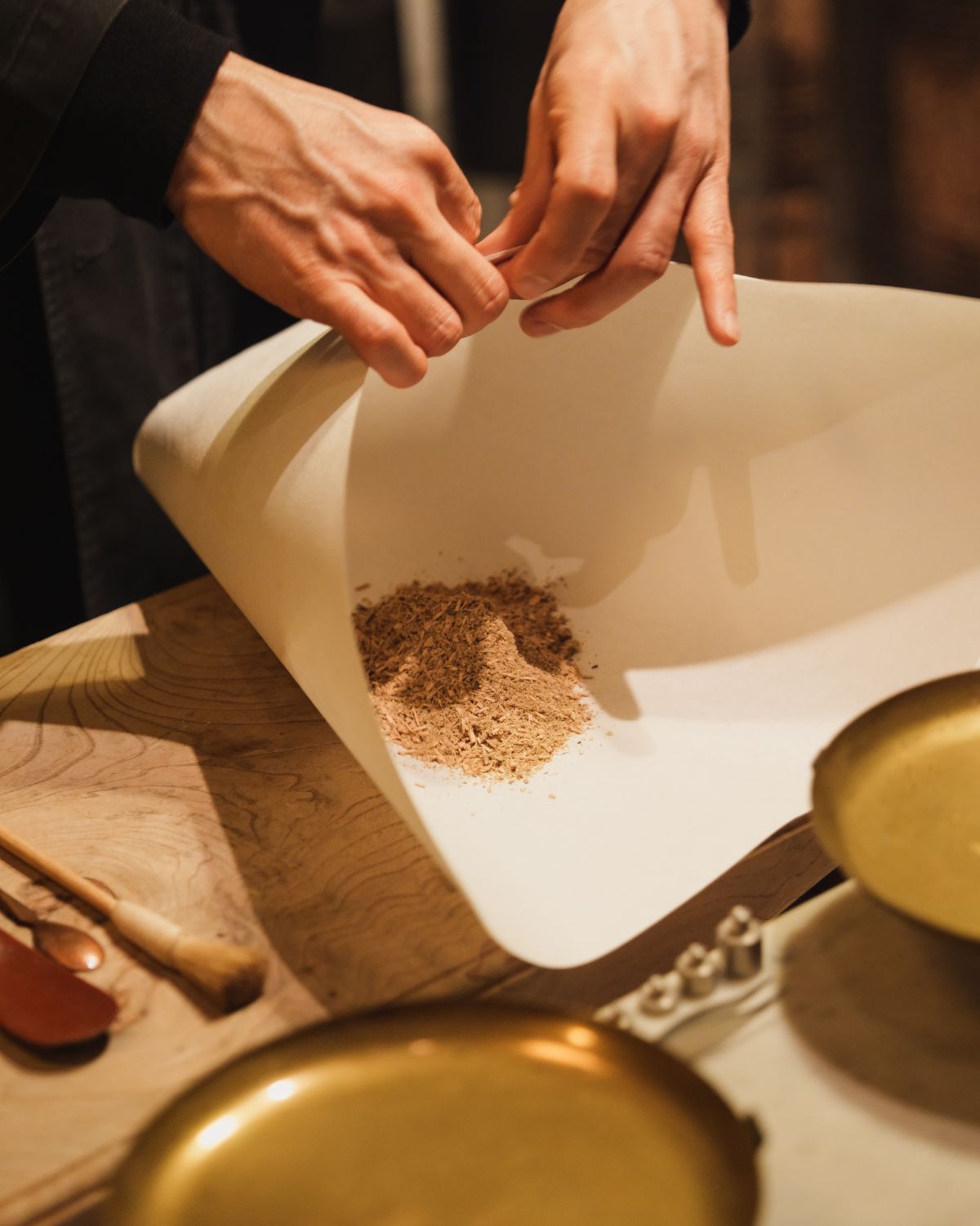 A black dressed person wrapping tea inside paper, on top of a wood table, with brushes on the side and golden bowls in front.