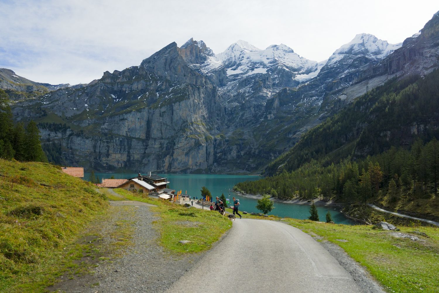A snowy and rocky mountain, behind a turquoise lake, in front of a road that concludes in wooden cabins, where people walk with backpacks.