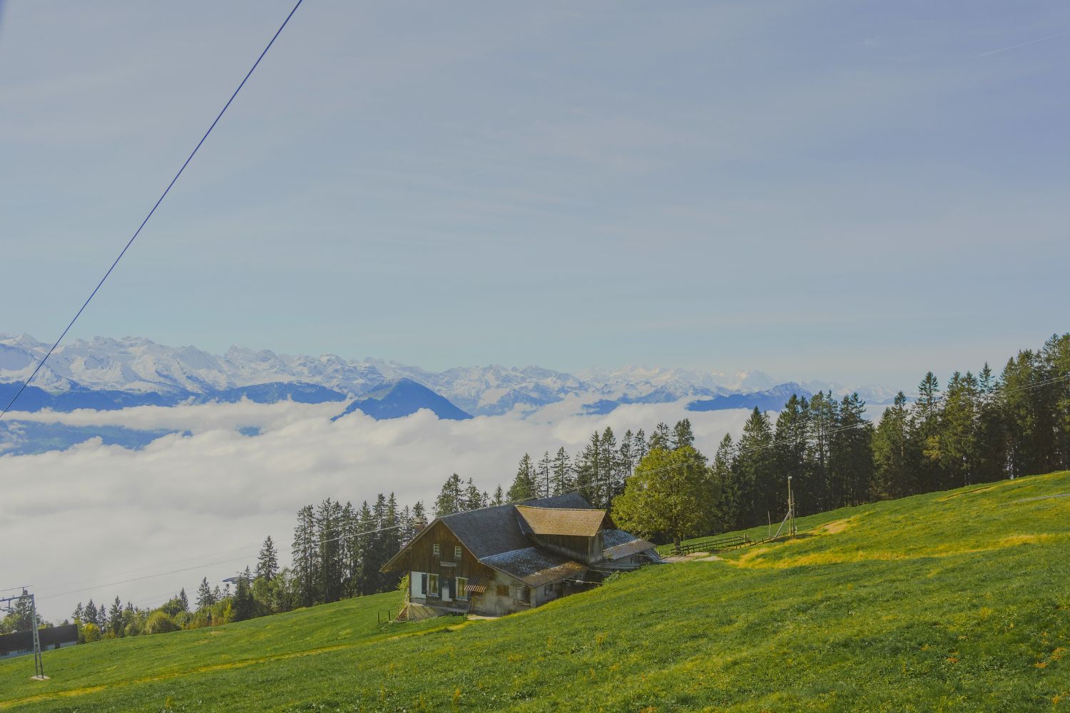 Wood cabin on a green hilltop, overlooking an array of mountains peaking through white clouds.