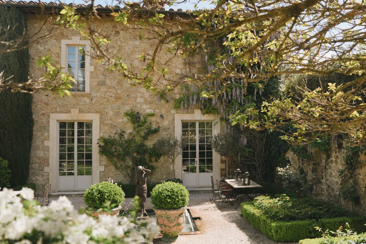 A cobbled stone house, with white french doors and wisteria, surrounded by a tailored garden.