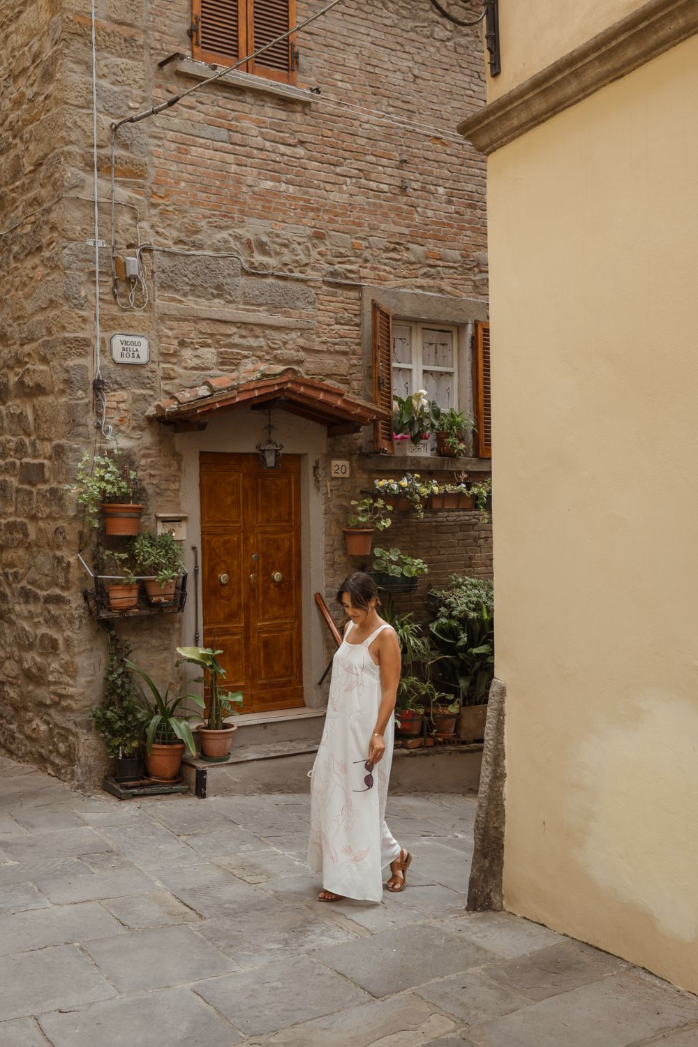 Woman in a white dress, standing in a street with a cobbled stone house, with wooden shutters and a door, next to hanging plants in vases.