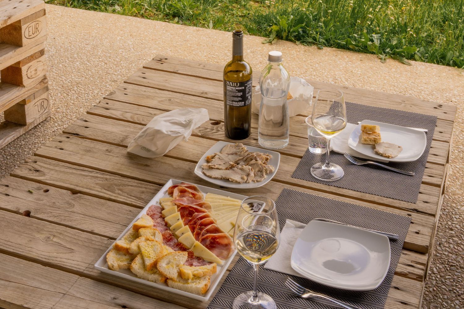 Center wooden table with cutlery, glasses, cheese, wine, water, bread and cured meats.