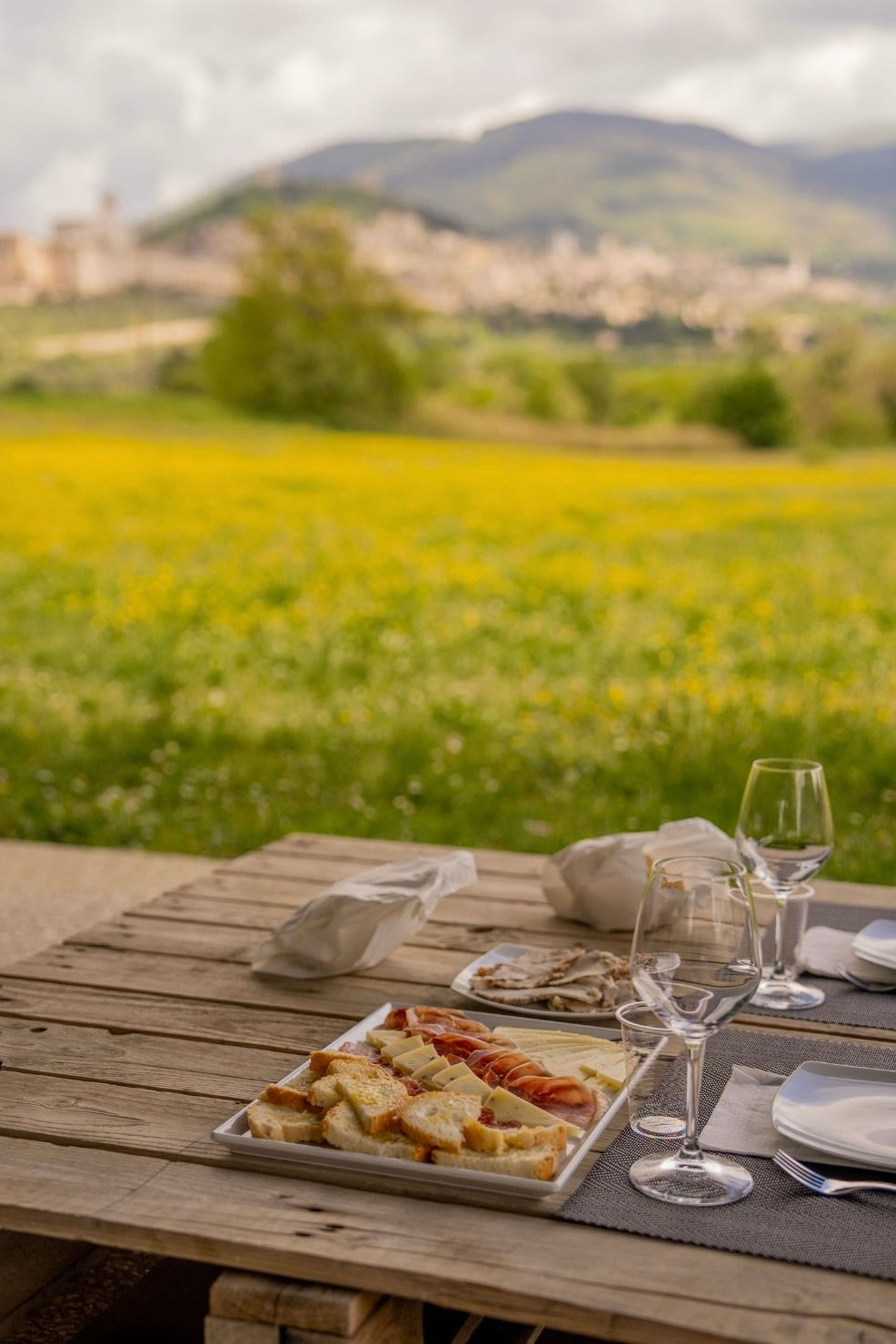 Center wooden table with cutlery, glasses, cheese, bread and cured meats, with a golden picturesque village amidst a green valley and vines in the background.