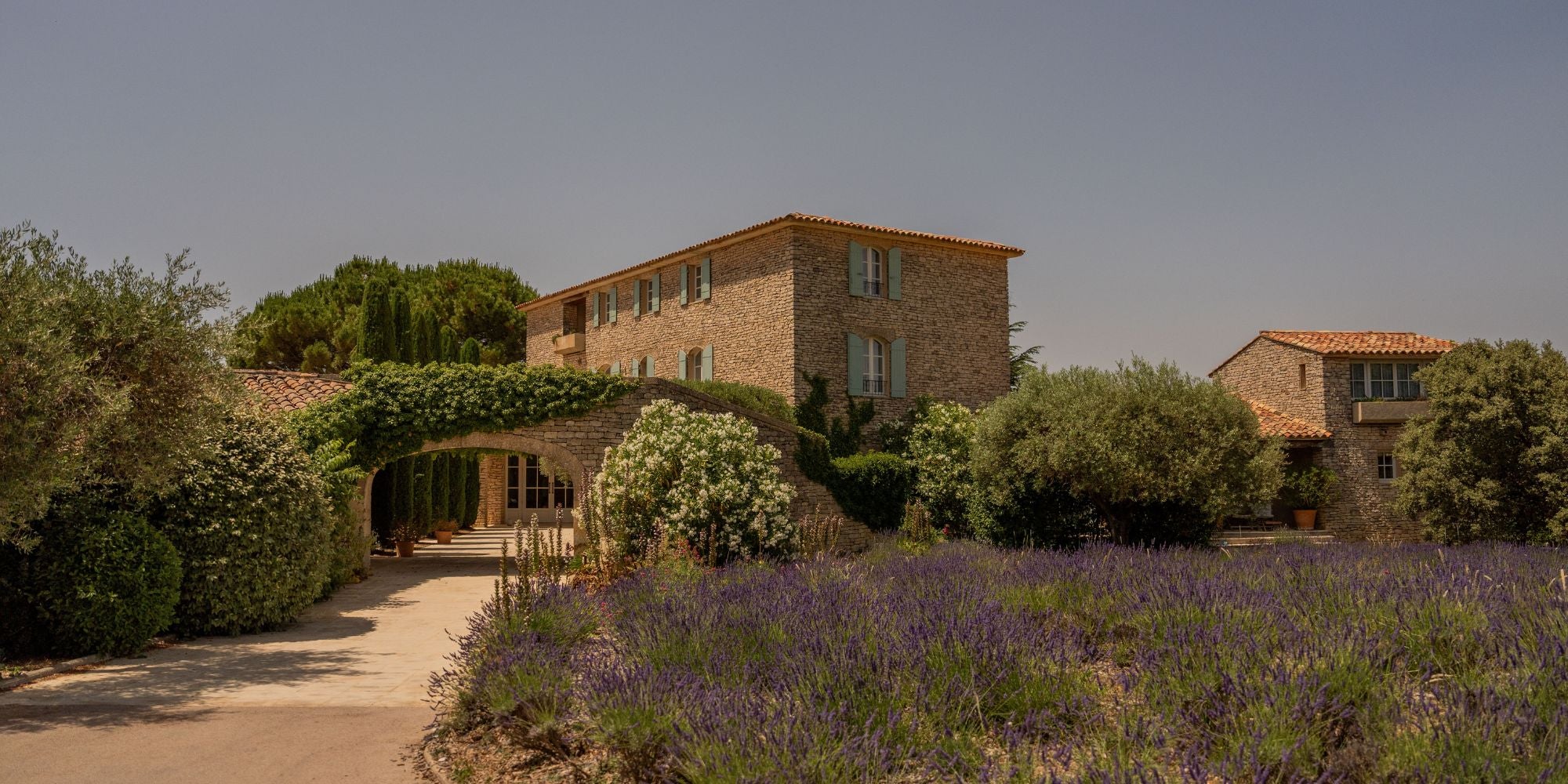 Cobblestone house, with light blue shutters, an archway with climbing trees, an abundant garden in front with lavender bushes.