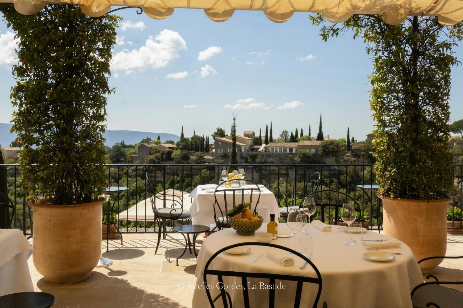 Restaurant balcony, with iron black chairs, white linen covered tables, large vases with climbing plants, and several houses in the background, amidst high trees.