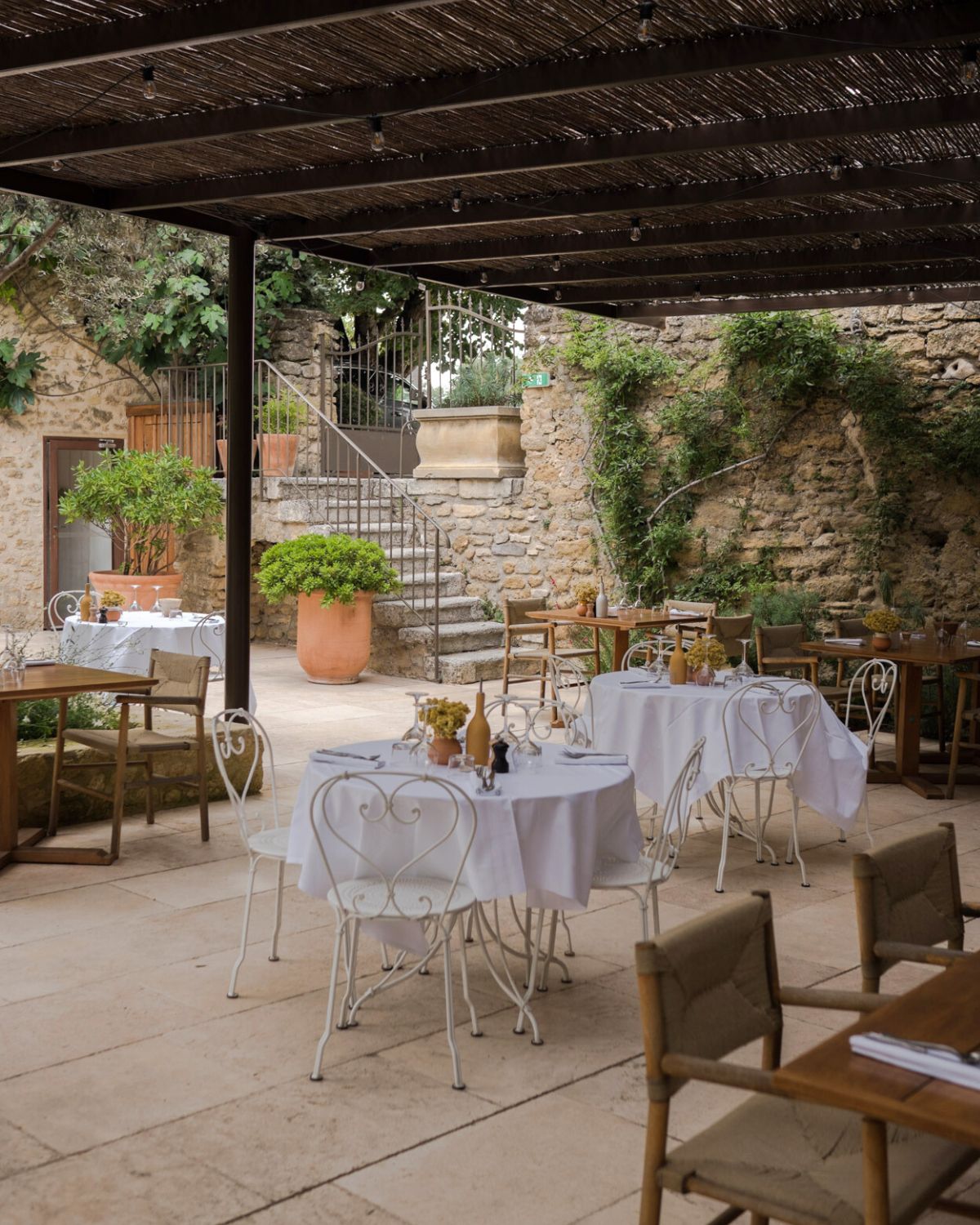 Outdoor restaurant setting, with iron white chairs, rounded white linen tables, a patched ceiling, a stone wall and vases with plants.
