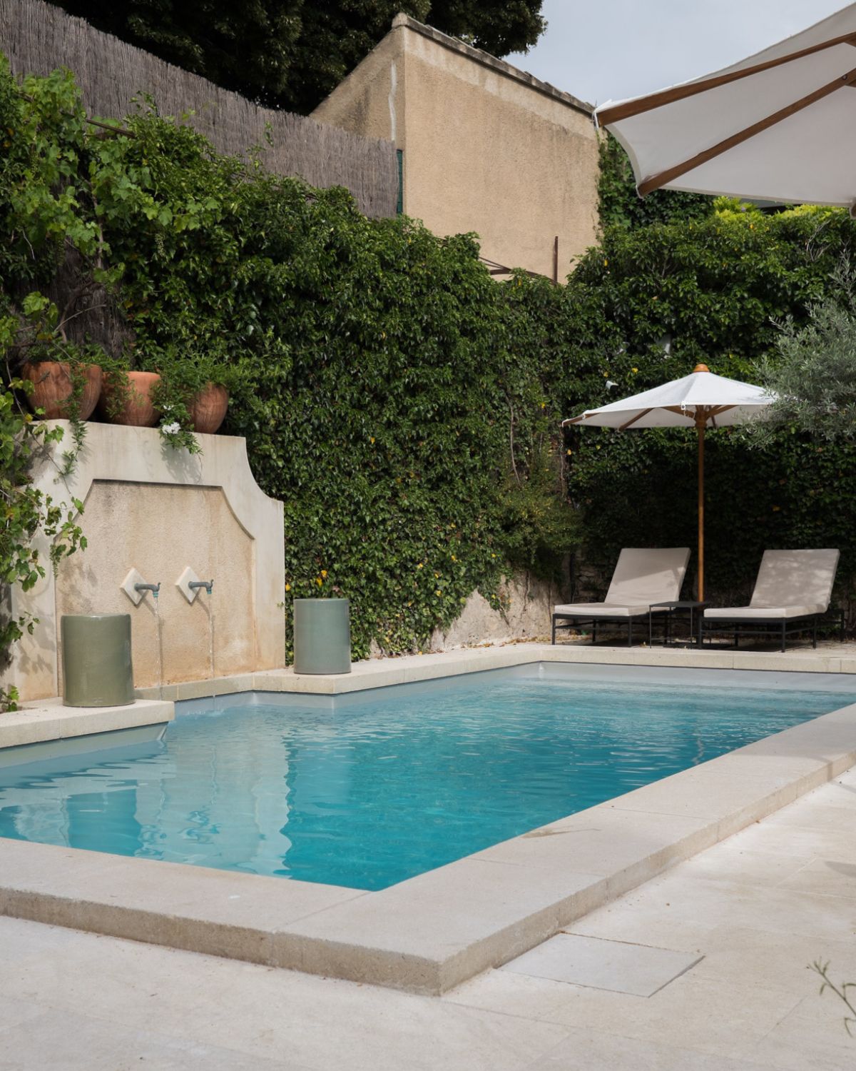 Pool with white sun beds , a white sunshade, a fountain and climbing plants on the walls.