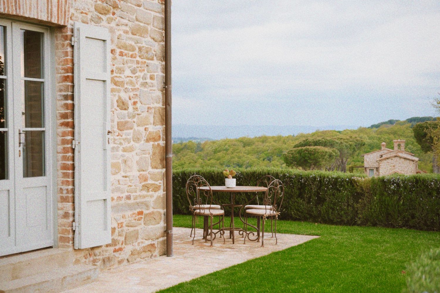 Iron outdoor set on the corner of a cobbled stone house, with light grey shutters, surrounded by a green scenery of trees, bushes, grass and a second cobbled stone house in the background.