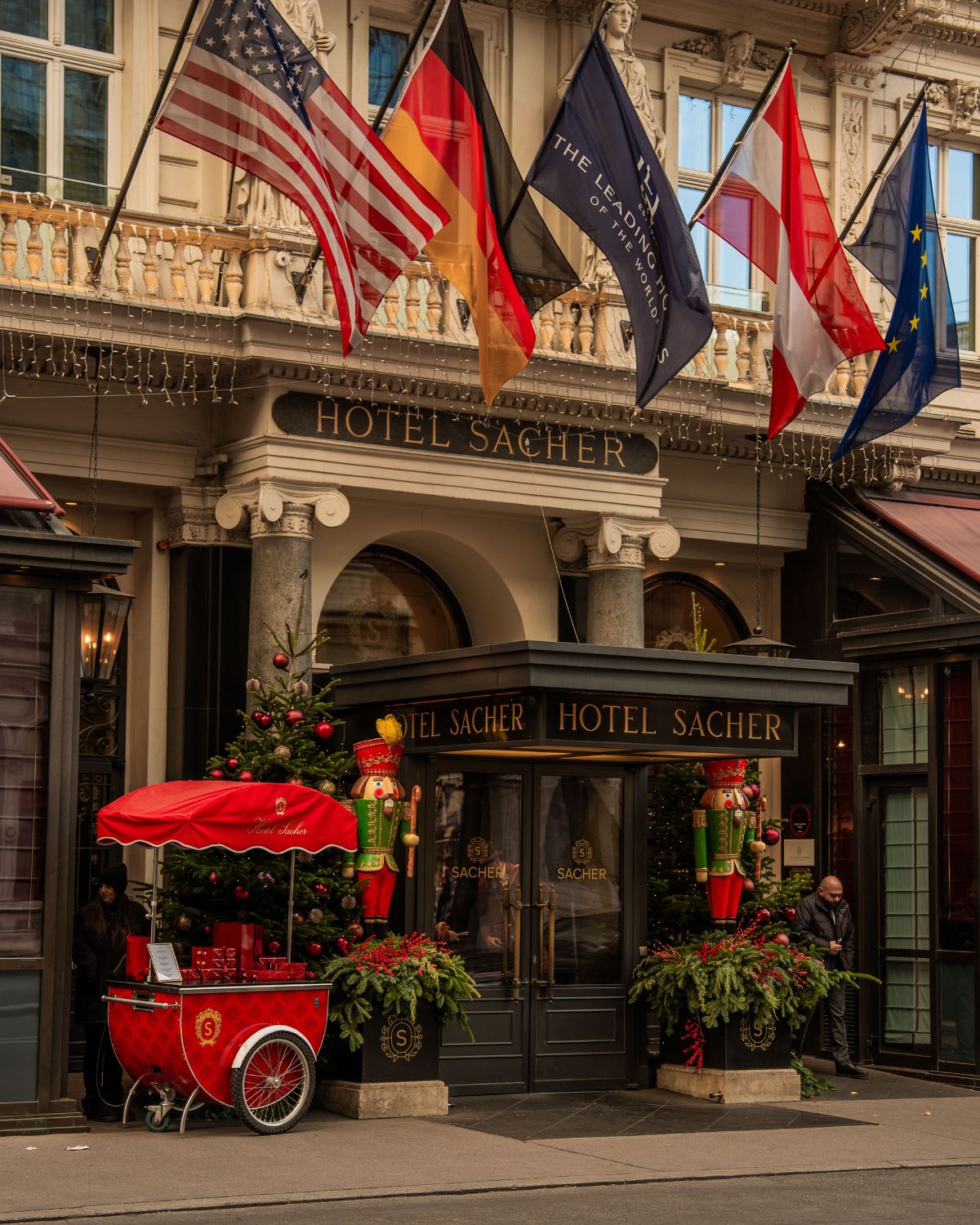 A classic hotel façade with a dark grey entrance door, Christmas ornaments and several flags on the top balcony.