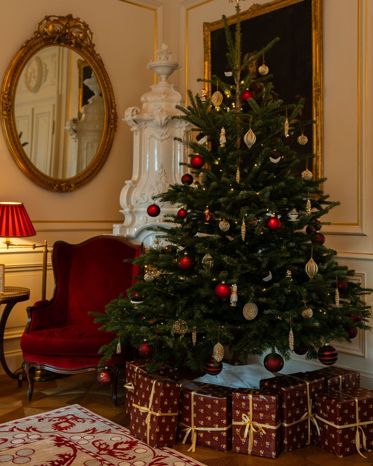 A hotel room, with gold paneling and ornamented mirror, an antique red armchair, next to a Christmas tree with red and gold ornaments, a red and white rug and a red standing lamp.