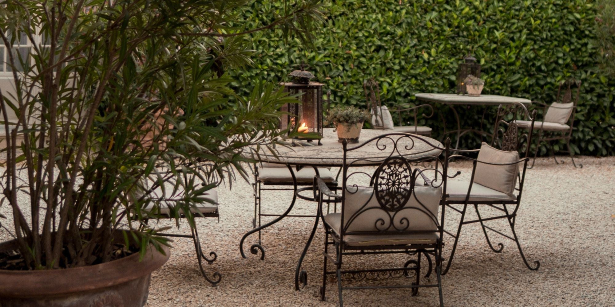 Dark iron garden furniture, with light cushions, a lit lantern on top of the table and a garden in the background.