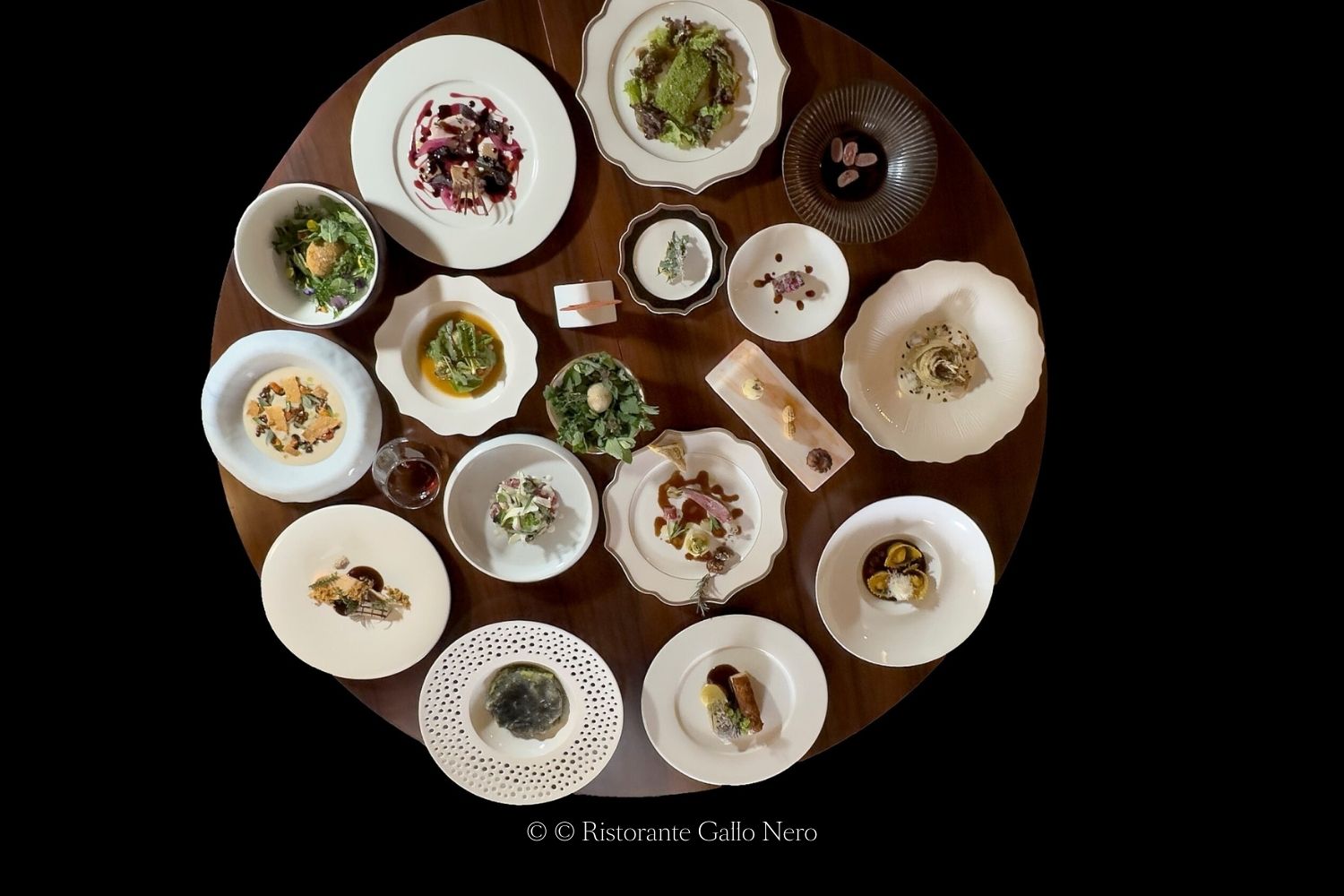Multiple white dishes with main courses and entrees, over a dark rounded wooden table over a black background.