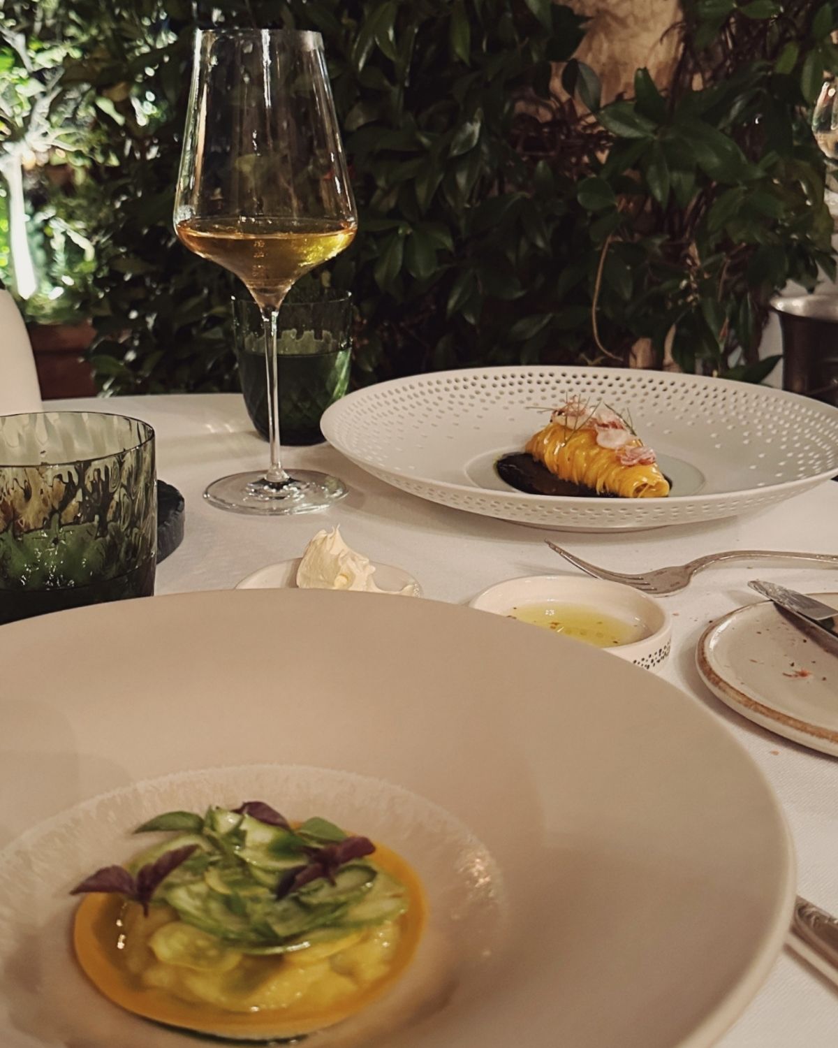 White dishes with well plated, small portioned food. A glass of white wine on a whit tablecloth in front of a green bush.