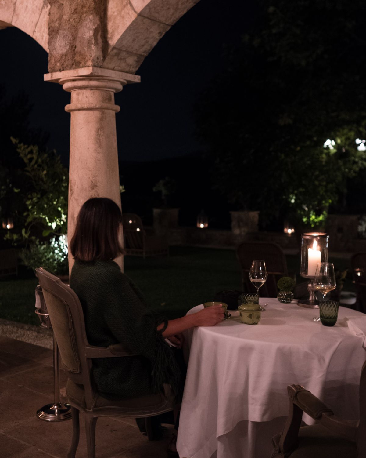 Restaurant setting at night. Round table with white linens, under arches, in front of a dark Tuscan countryside landscape and a woman in a dark dress.