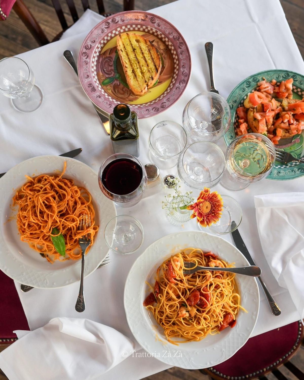 Four different dishes, with Italian food on them, white linens and red and dark wooden chairs.