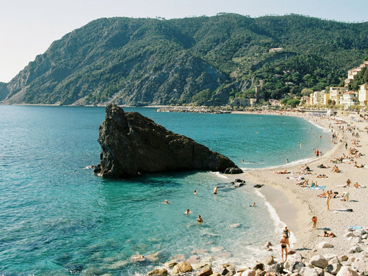 The beach of Monterosso al Mare, in Cinque Terre, with its crystal clear waters, the green hills on the background, and its iconic Scoglio di Monterosso in the middle.