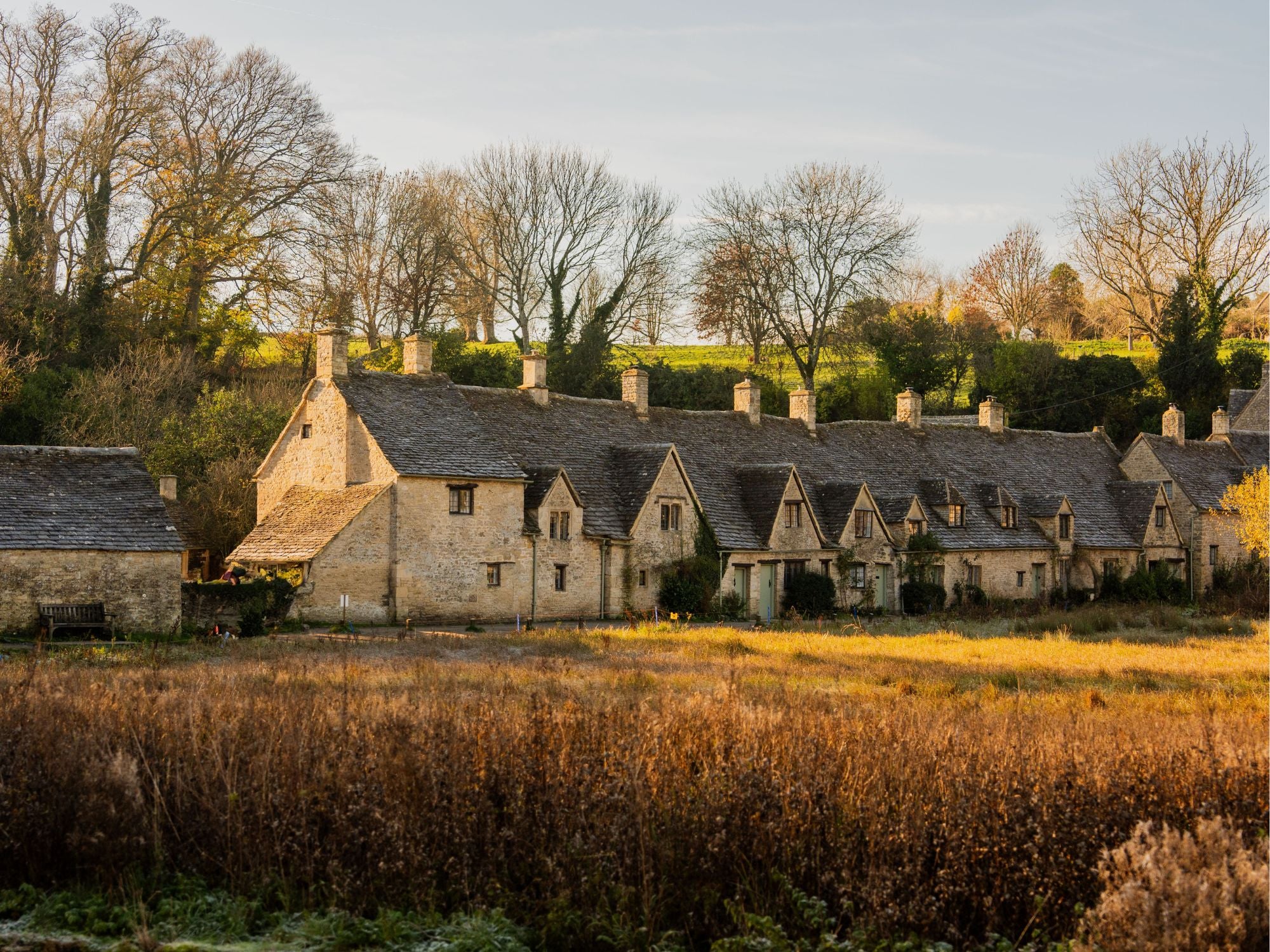 The row of stone cottages at Arlington Row, in the village of Bibury, Cotswolds, surrounded by green rolling hills and meadows.