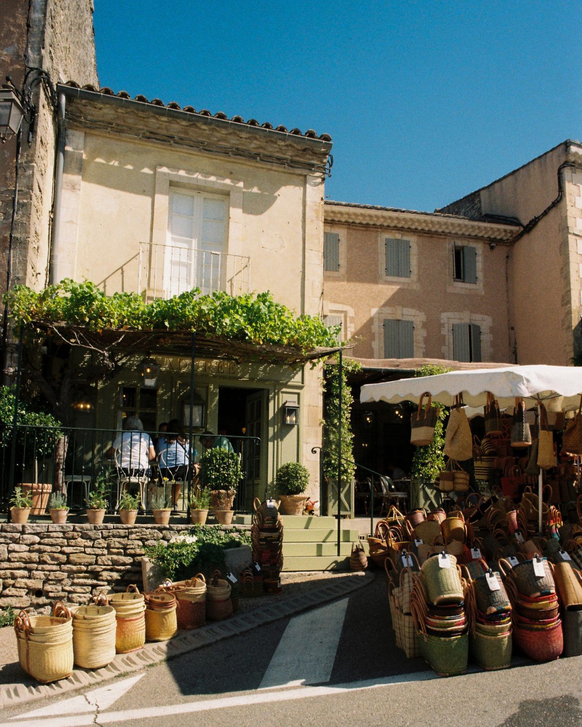 Street in Gordes with ligh blue shutters, a Ladurée location, outdoor iron sitting and market baskets.