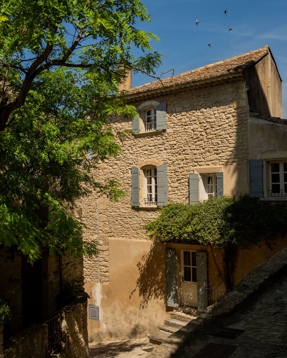 A cobblestone house with light-blue shuttered windows and four birds flying.