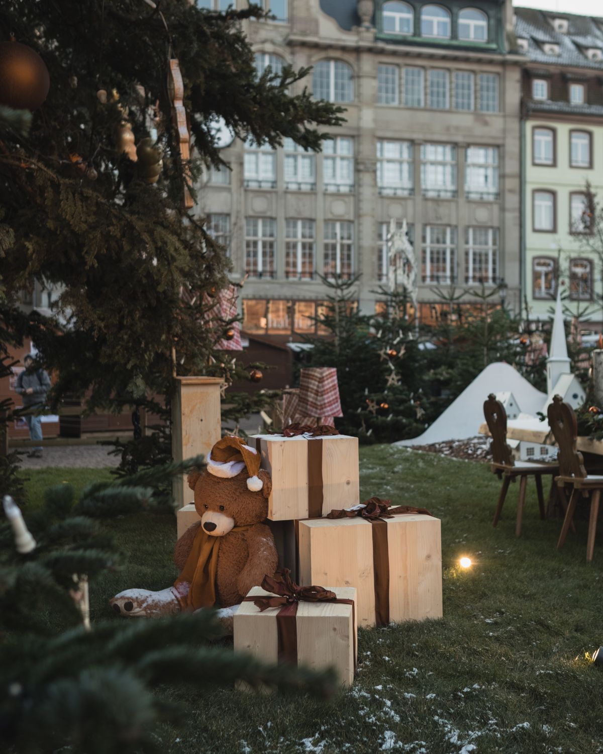 Christmas market outdoor setting. A brown bear sitting next to wooden gifts, next to a Christmas tree, facing the city's buildings.