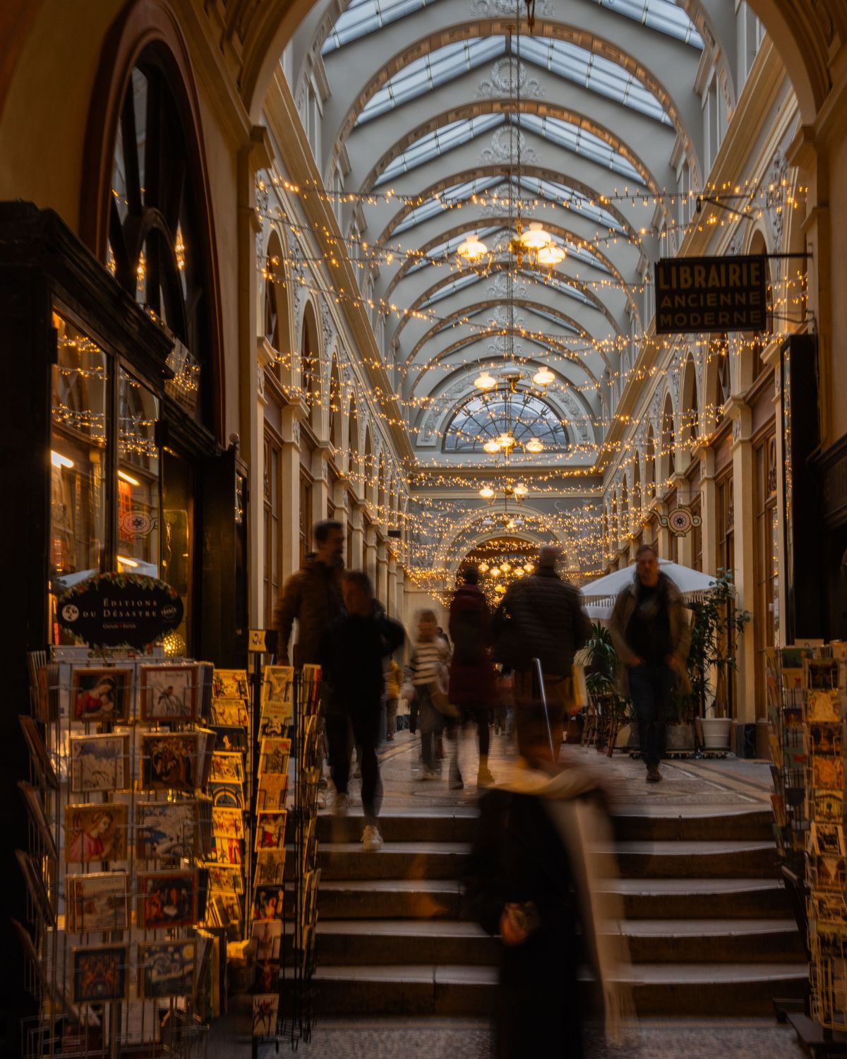Hallway with Christmas lights and glass ceilings above archways, over people walking pass stores.