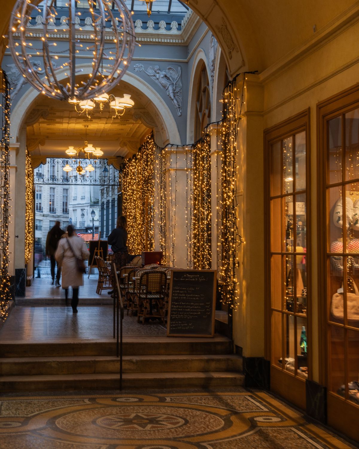 A corner of an interior hall with Christmas lights, glass ceilings, classic buildings outside, and wooden store windows.