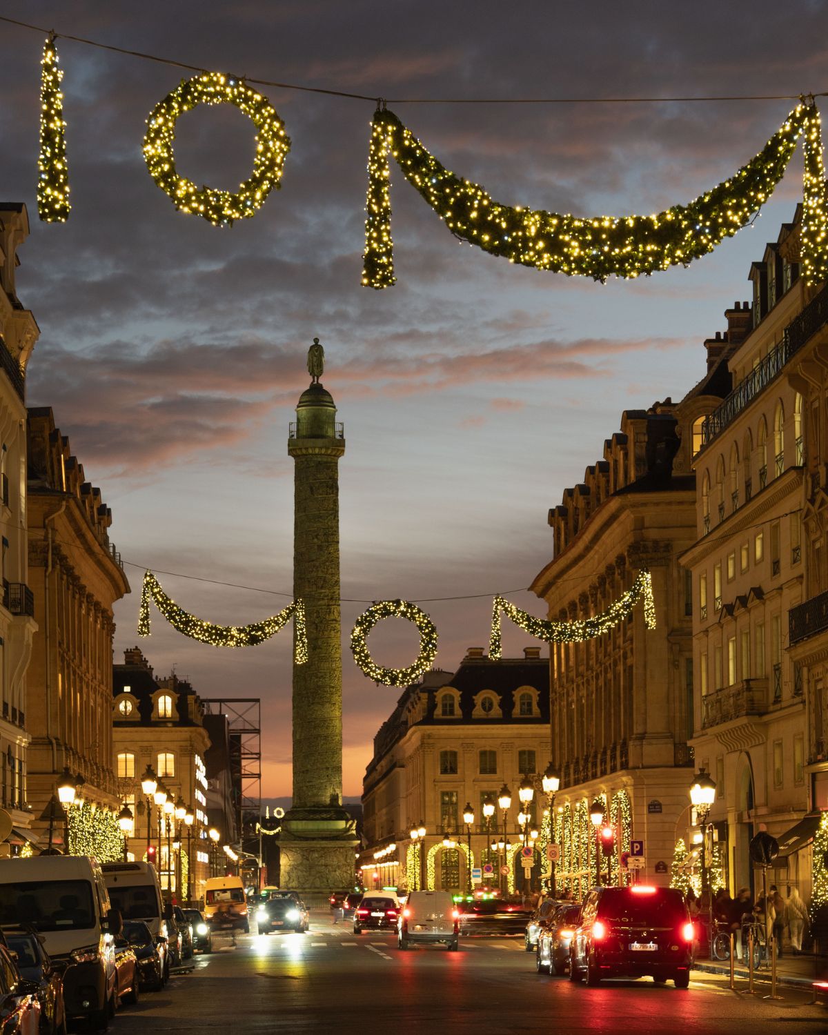 Paris square at sunset, with classic buildings and a tower at the middle of the street where cars pass through. Christmas ornaments adorn the streets, and the doors and windows of the buildings.