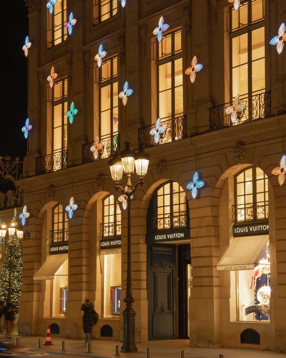 Classic building with the store Louis Vuitton logo and emblems adorning the façade, next to a Christmas tree and behind an antique black outdoor lamp.