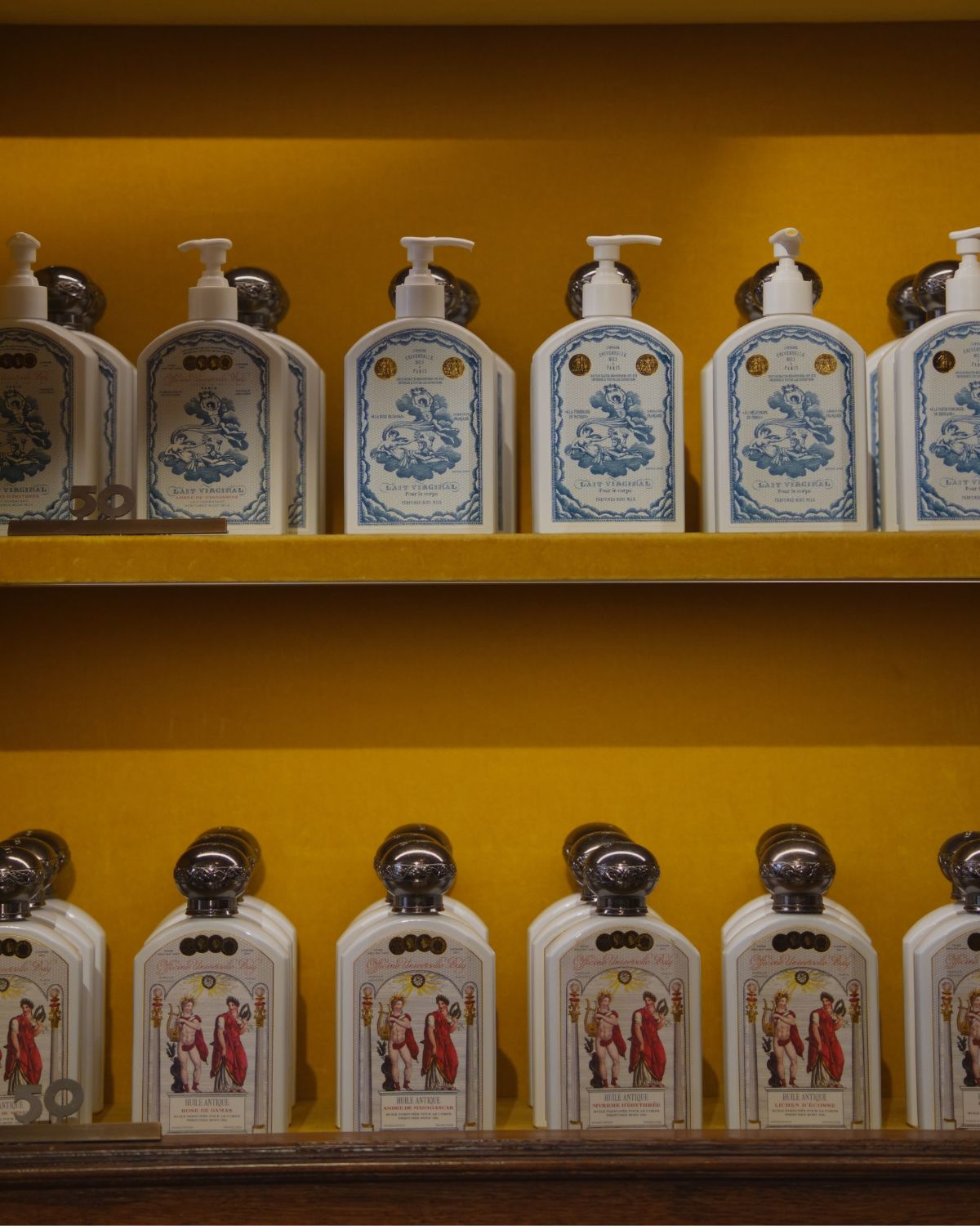 Red and blue antique like bottles of beauty products, in front of a velvet yellow wall, on a wooden shelving.