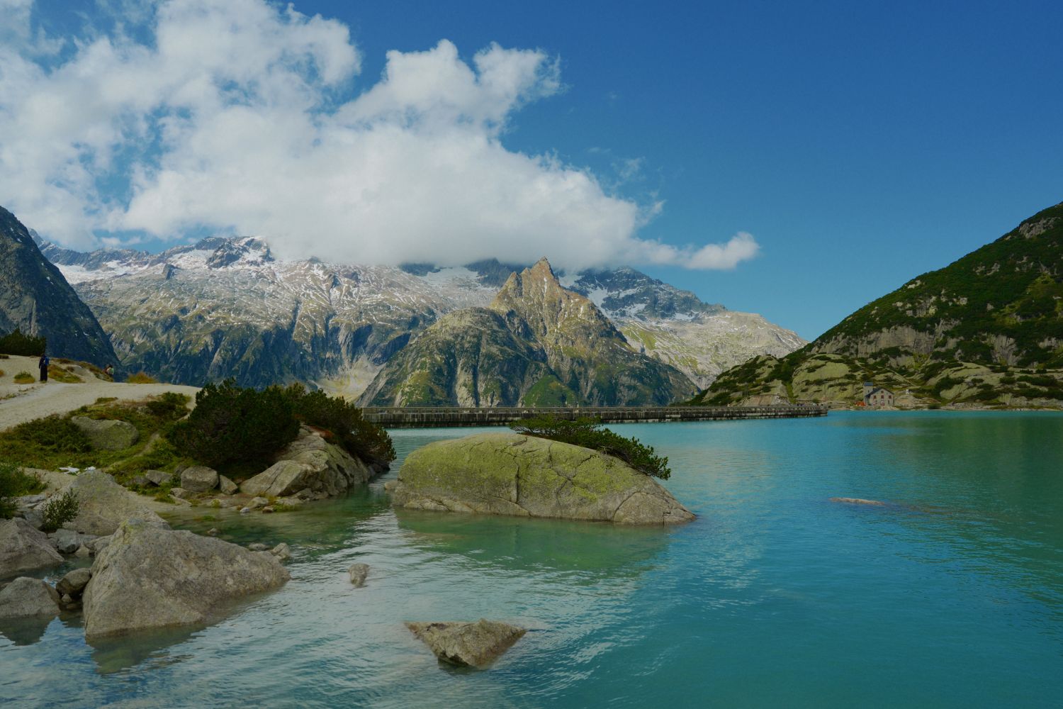 Turquoise lake amidst green rocky and snowy mountains.