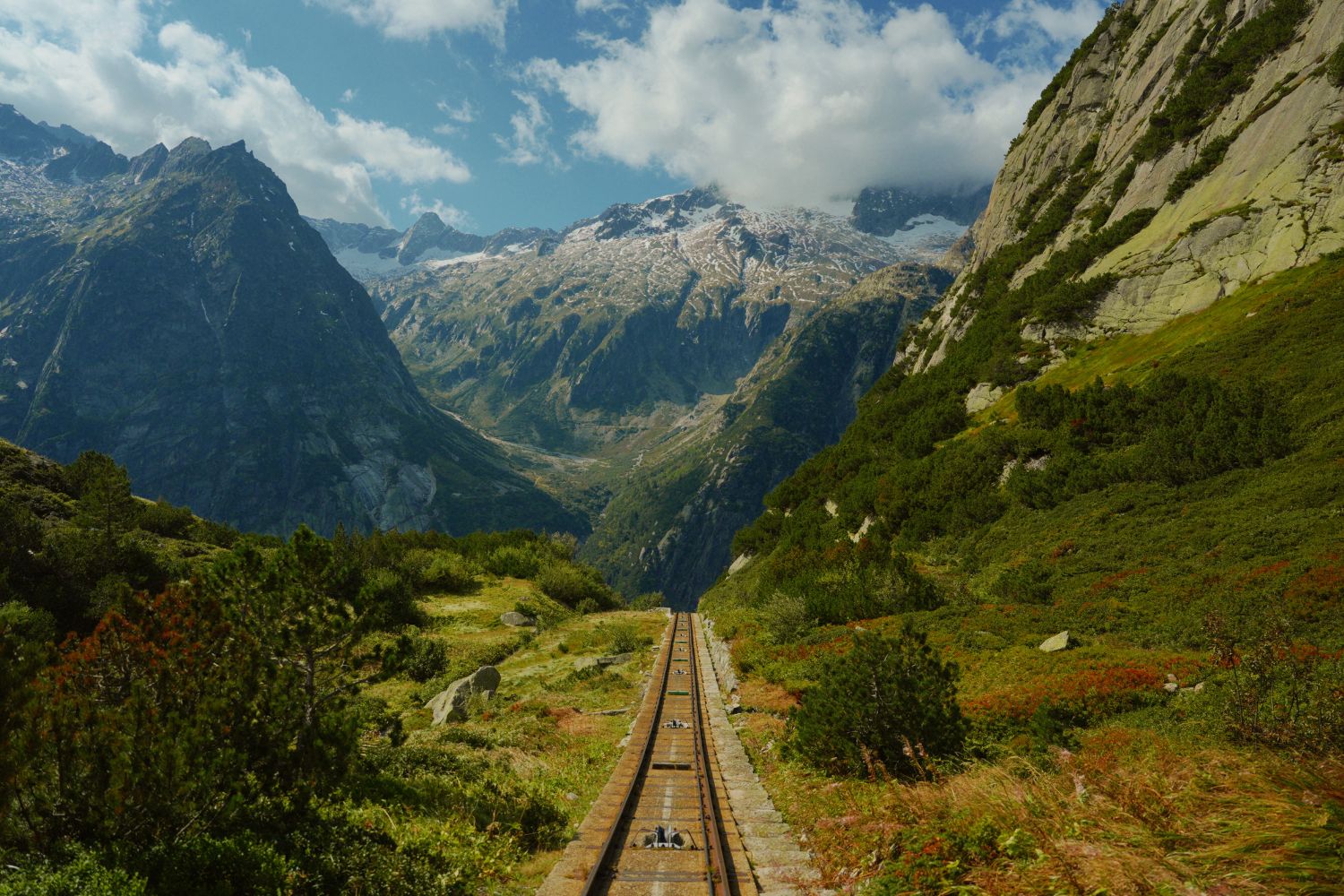A train rail, in the middle of green, rocky and snowy mountains.