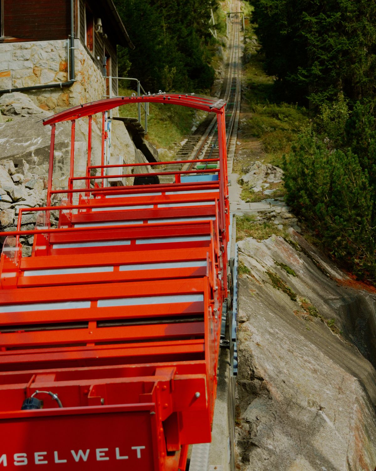 A red train, going up a rail on a hilltop green mountain.