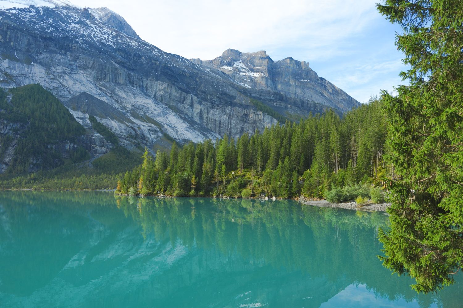 A rocky and snowy mountain behind a turquoise lake, with luscious green trees on the side, next to a rocky shore.