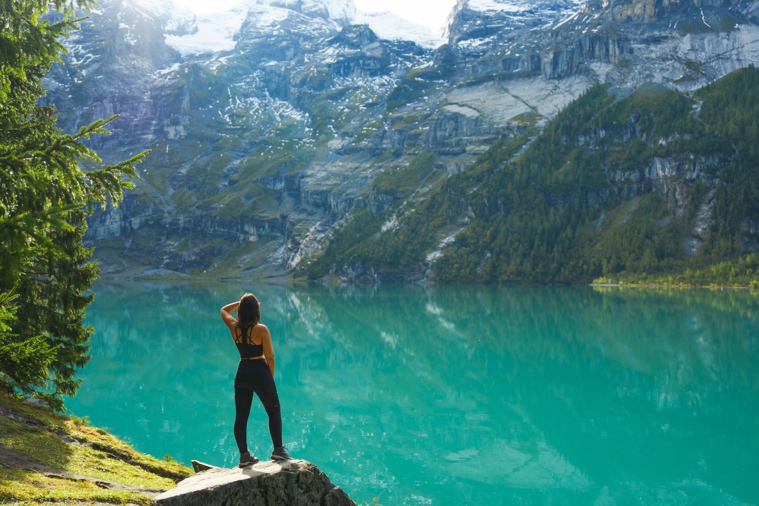 A lady with black sportswear, on top of a rock, facing a turquoise lake with rocky and snowy mountains behind.