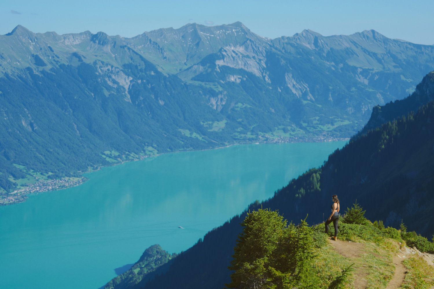 A lady with black sportswear, on top of a green mountain, facing a turquoise lake with rocky and green mountains.