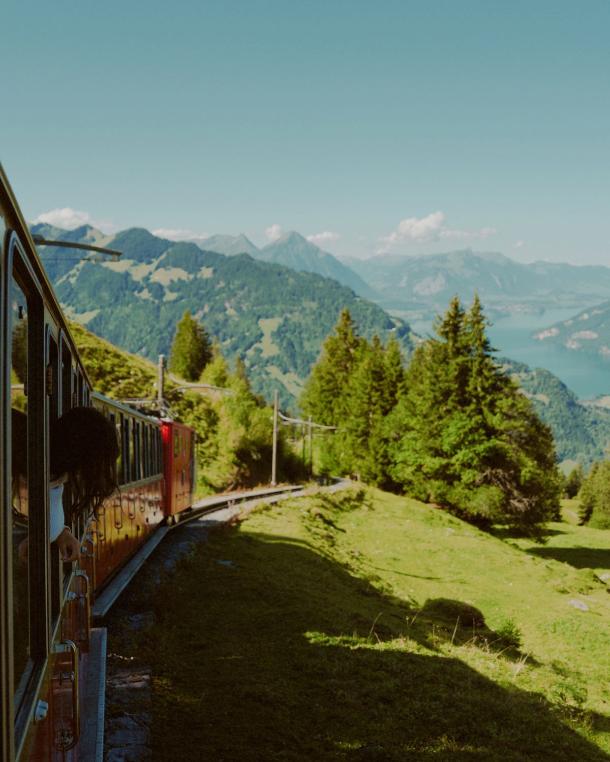 A woman on the window of a red train, crossing a light green valley, with dark green mountains in the back.