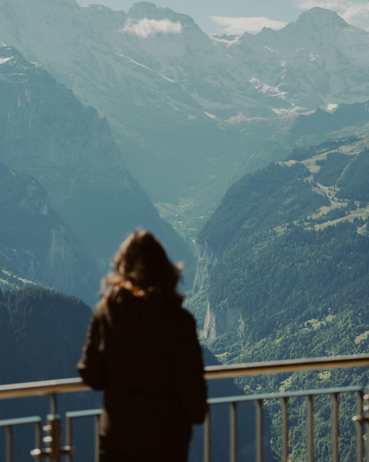 A lady with black sportswear, behind a fence, on a hilltop facing a an array of rocky mountains.