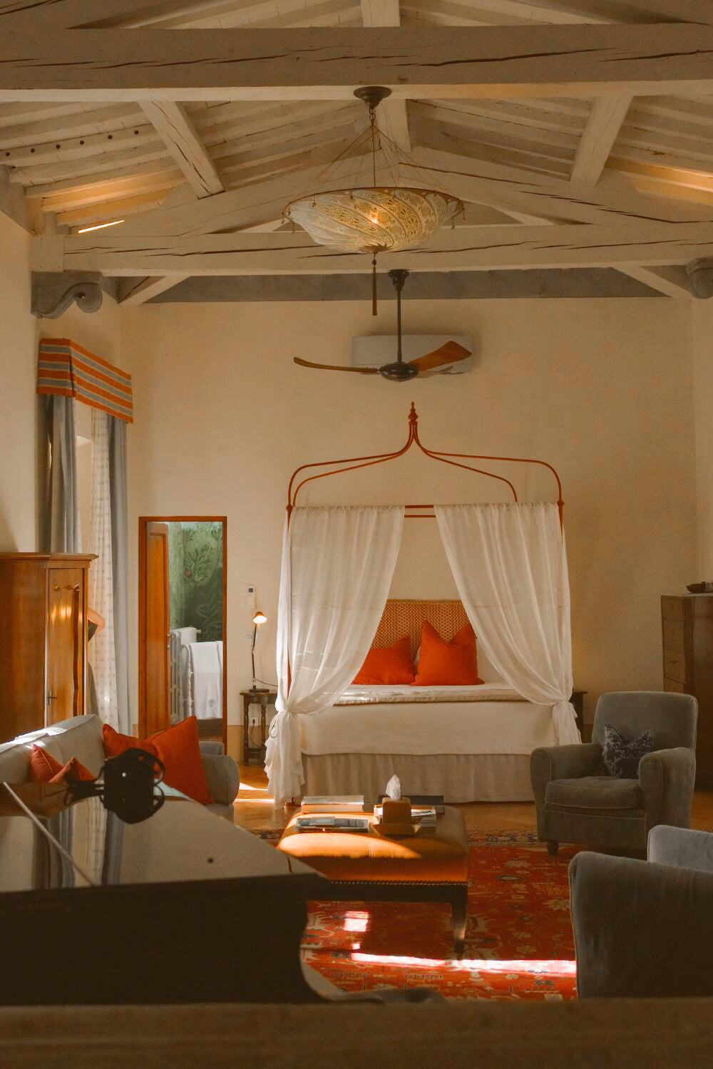 Hotel room with a white linen bed and curtains, red pillows, a warm toned living room with a piano, blue armchairs and white wooden beams on the ceiling.