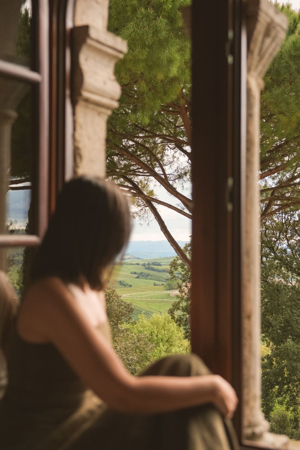 Young lady with a dark green dress, sitting on a window balcony, facing the green Tuscan hills.