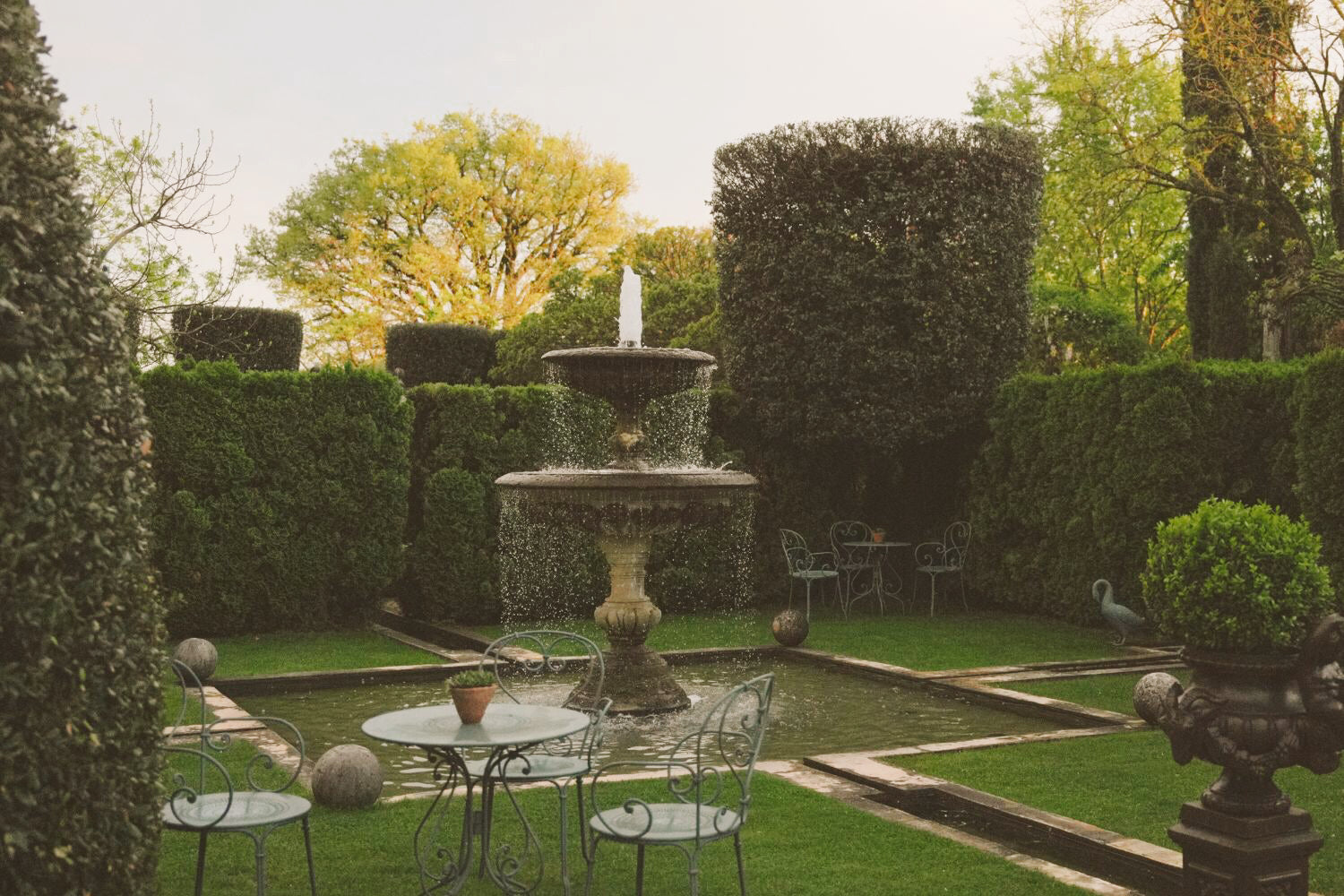 A tailored garden, with carved bushes, a fountain and green ironed outdoor chairs and tables.